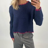 En person iført en mørkeblå sweater med pink kant detaljer holder en smartphone i hånden. Baggrunden er neutral, og personen bærer grå bukser og har lyst hår.