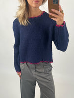 En person iført en mørkeblå sweater med pink kant detaljer holder en smartphone i hånden. Baggrunden er neutral, og personen bærer grå bukser og har lyst hår.