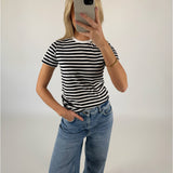 En person iført sort-hvid stribet T-shirt og blå jeans tager et spejlbillede med en smartphone i et minimalistisk rum.