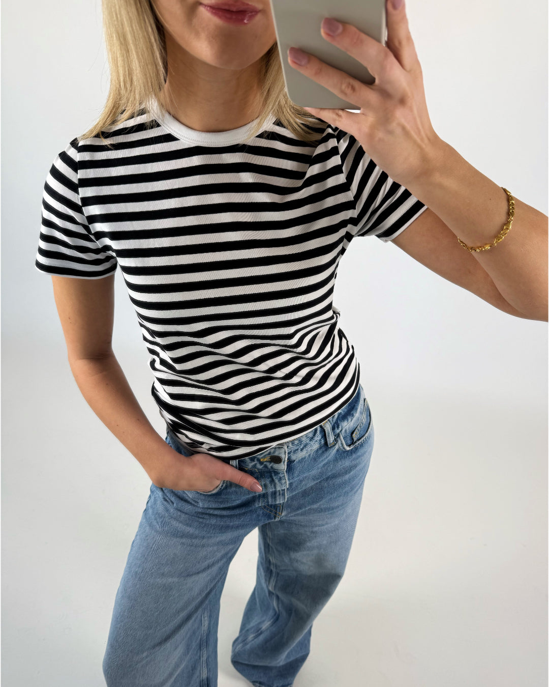 En person iført en sort-hvidstribet t-shirt og blå jeans tager en selfie. De står i et lysegråt rum og har en hånd i bukselommen.