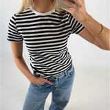 En person iført en sort-hvidstribet t-shirt og blå jeans tager en selfie. De står i et lysegråt rum og har en hånd i bukselommen.