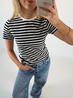 En person iført en sort-hvidstribet t-shirt og blå jeans tager en selfie. De står i et lysegråt rum og har en hånd i bukselommen.