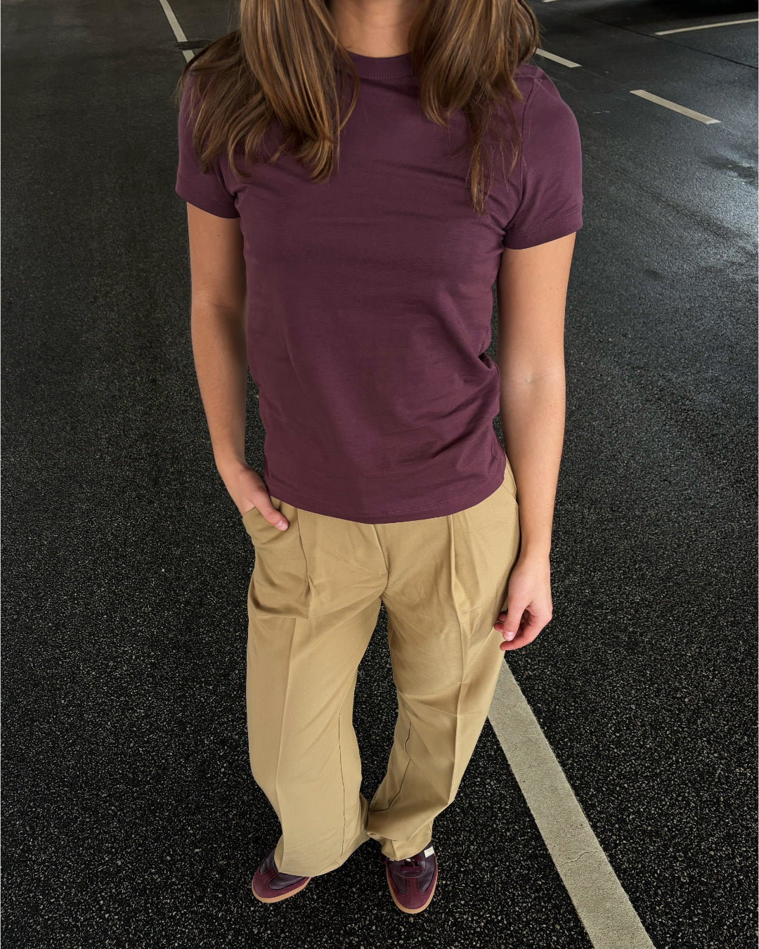 Person står på en sort asfaltvej. De har en lilla t-shirt og beige bukser på. Hånden er i lommen, og de bærer brune sko. Vejen har hvide markeringer.