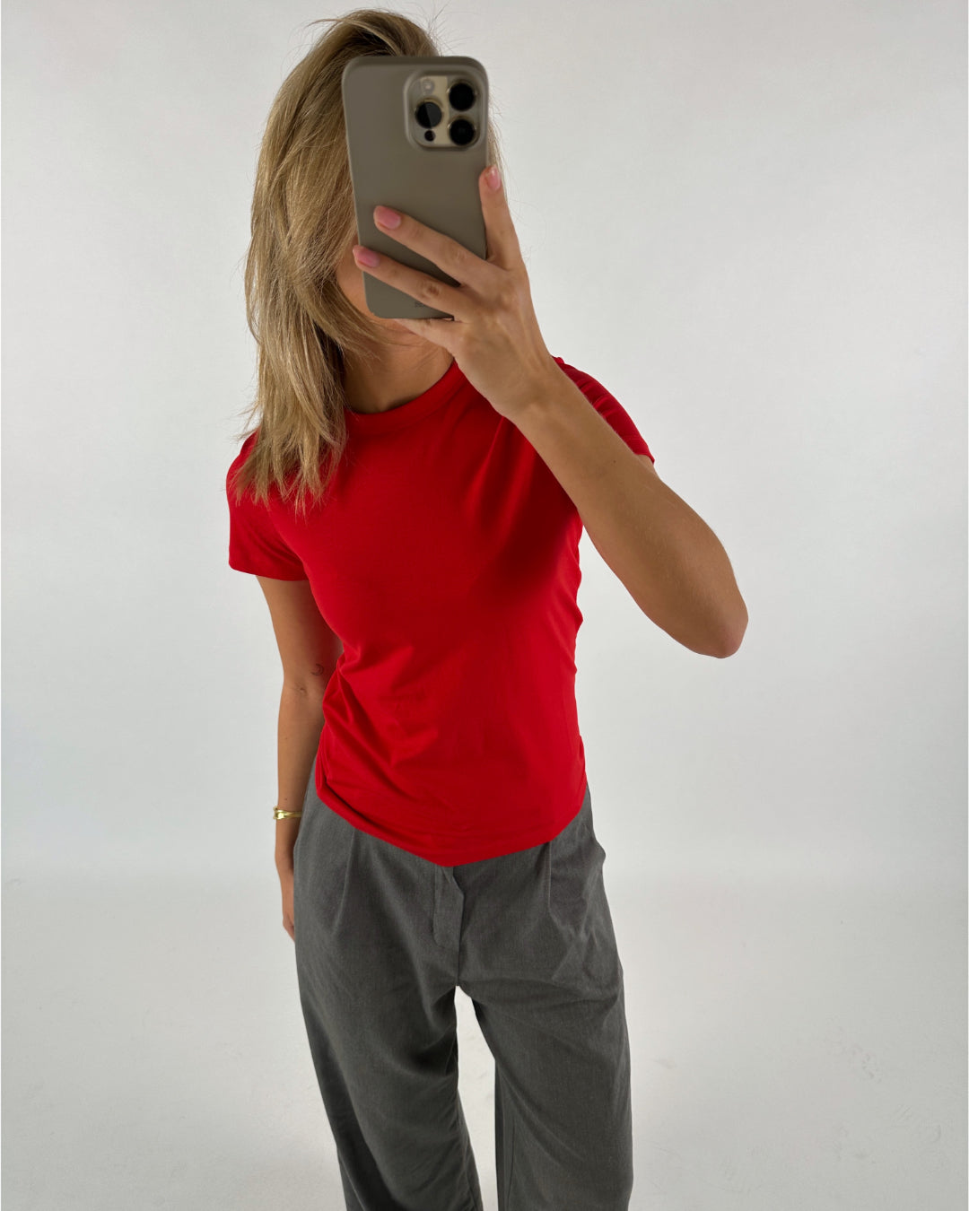 Person i rød t-shirt holder smartphone for ansigtet, står foran hvid baggrund. Personen bærer også grå bukser og har langt blondt hår.