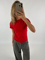 Person i rød t-shirt holder smartphone for ansigtet, står foran hvid baggrund. Personen bærer også grå bukser og har langt blondt hår.