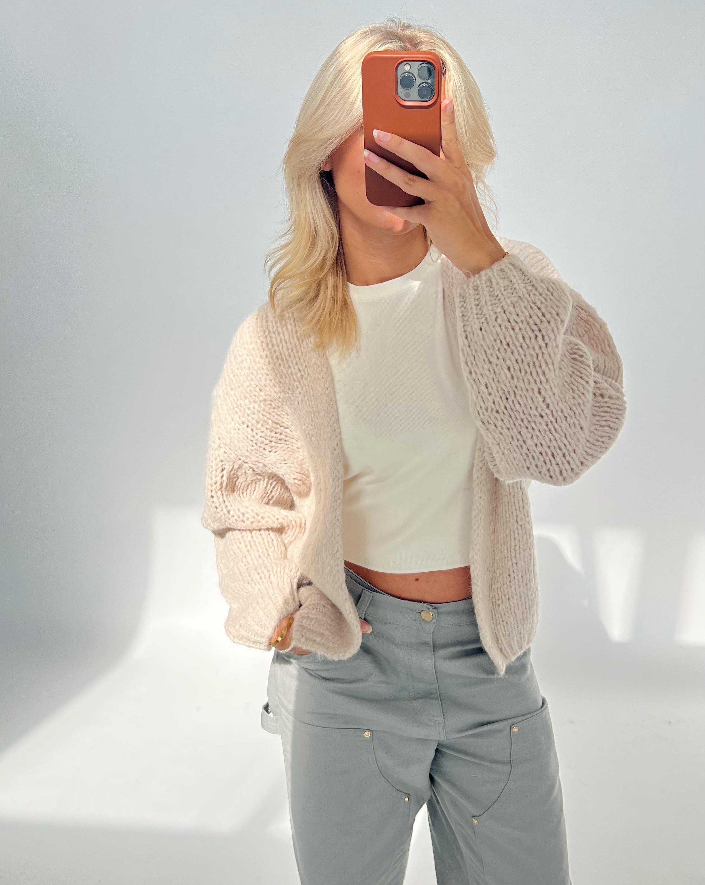 En person tager et selfie med en smartphone iført en lys striktrøje og grå bukser i et lyst rum. Ansigtet er dækket af telefonen.