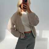 En person tager en selfie i et lyst rum iført en lys, strikket cardigan og grå bukser, og holder en smartphone op, der dækker ansigtet.