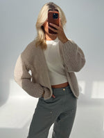 En person tager en selfie i et lyst rum iført en lys, strikket cardigan og grå bukser, og holder en smartphone op, der dækker ansigtet.
