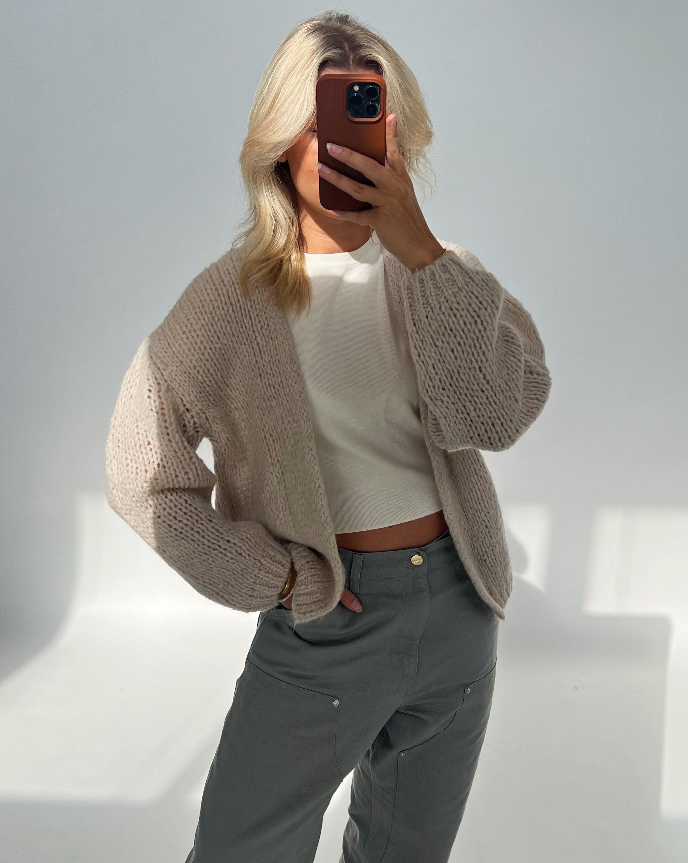 En person tager en selfie i et lyst rum iført en lys, strikket cardigan og grå bukser, og holder en smartphone op, der dækker ansigtet.