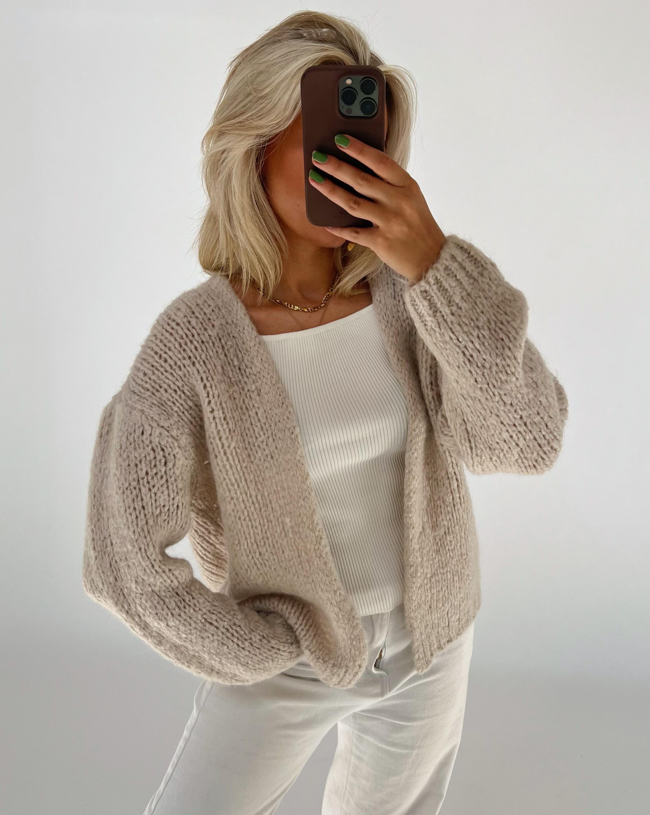 Person holder en mobiltelefon, iført en beige striktrøje over en hvid ribbet top. Grøn neglelak er synlig på neglene, og baggrunden er lys.