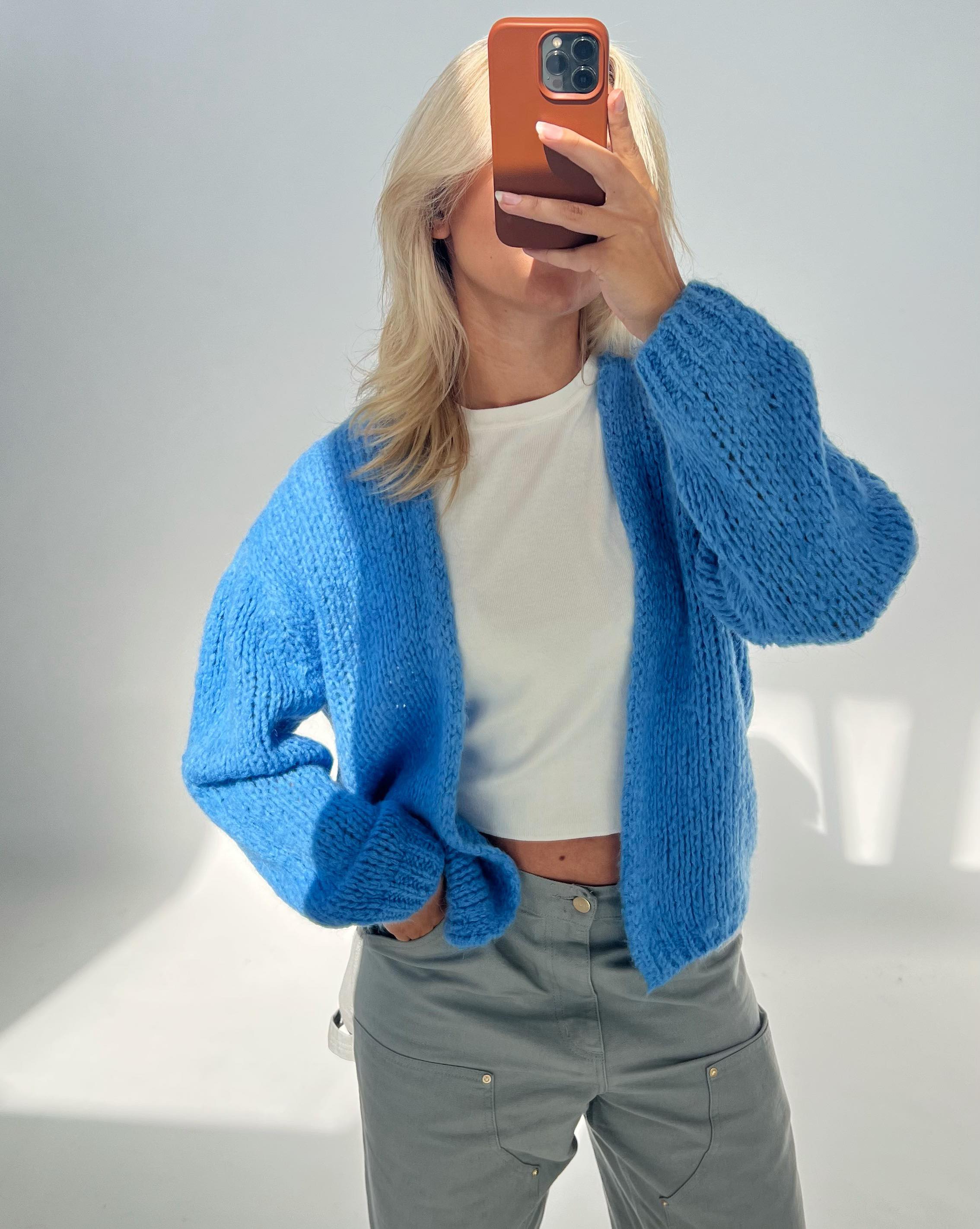 En person iført en lyseblå striktrøje tager et spejlbillede med en smartphone. Personen bærer også en hvid crop-top og grå bukser i et lyst rum.