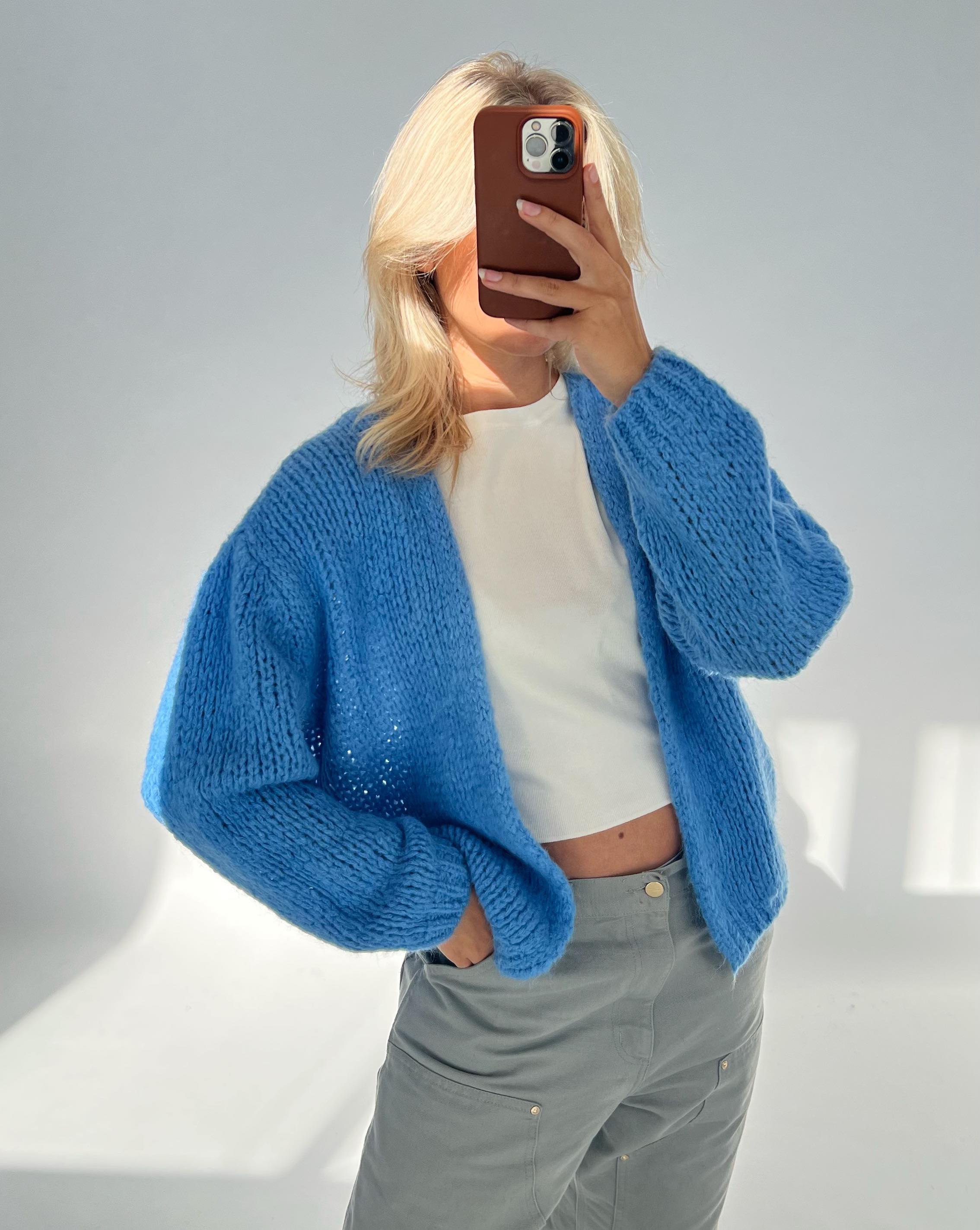 En person iført en blå strik cardigan og grå bukser holder en telefon op foran ansigtet i en lys, minimalistisk indstilling.