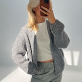 Person tager et spejlbillede iført en grå striktrøje og lyse bukser, dækker ansigtet med en mobiltelefon i et lyst, minimalistisk rum.