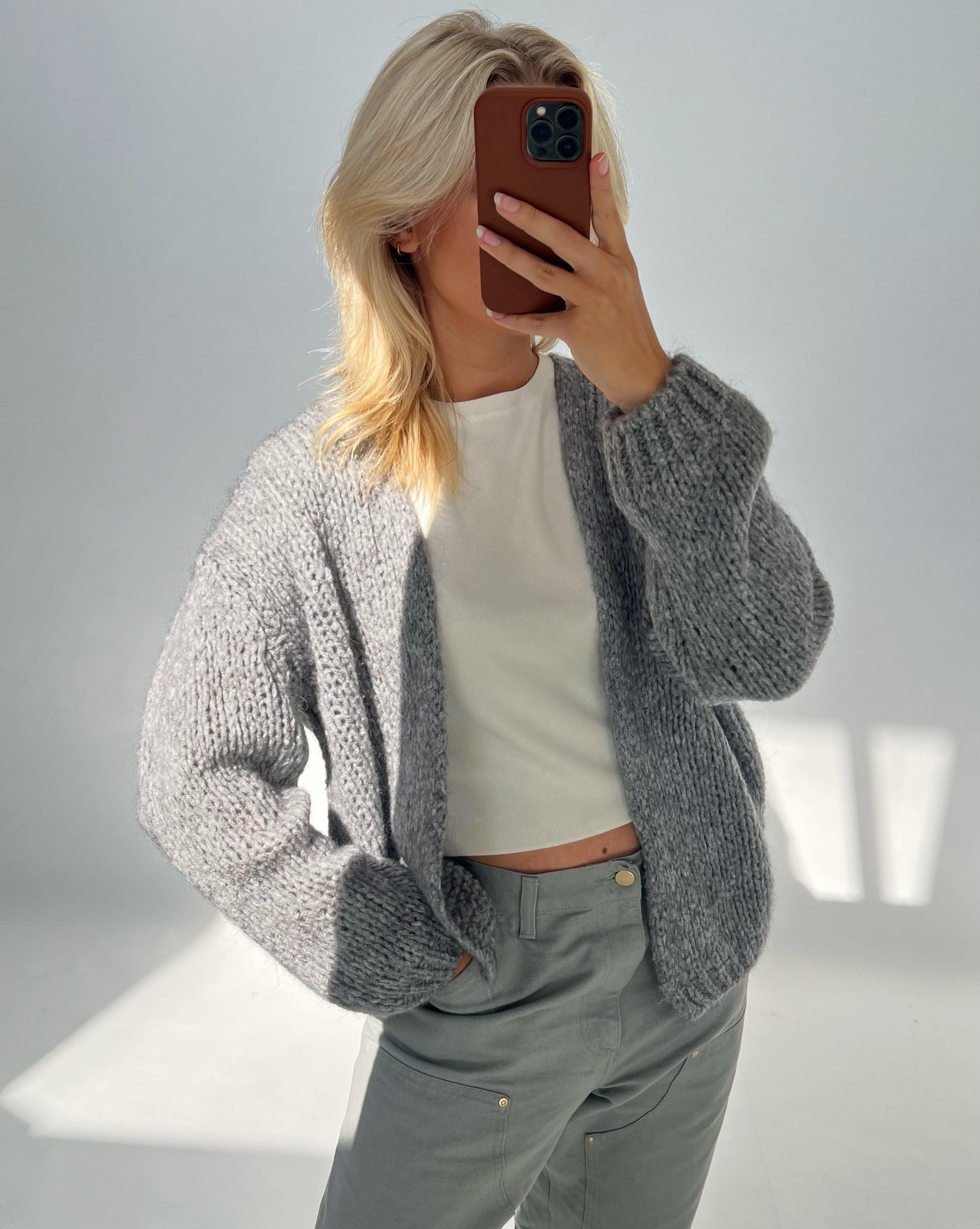 Person tager et spejlbillede iført en grå striktrøje og lyse bukser, dækker ansigtet med en mobiltelefon i et lyst, minimalistisk rum.