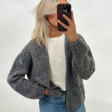 Kvinde i gråt strikket cardigan og hvid t-shirt tager selfie med telefon. Hun står i et lyst rum og har også jeans på.