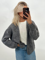 Kvinde i gråt strikket cardigan og hvid t-shirt tager selfie med telefon. Hun står i et lyst rum og har også jeans på.