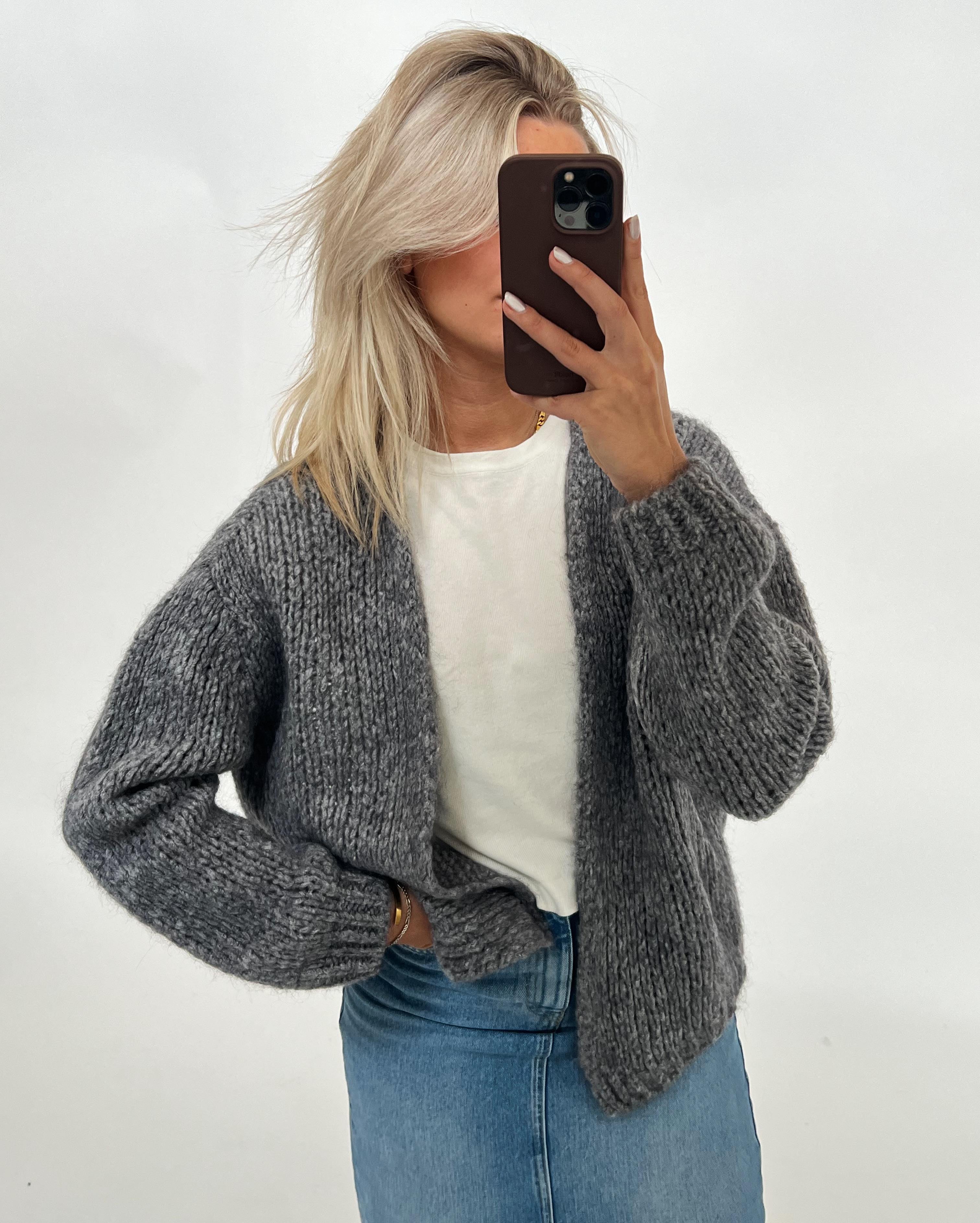 Kvinde i gråt strikket cardigan og hvid t-shirt tager selfie med telefon. Hun står i et lyst rum og har også jeans på.