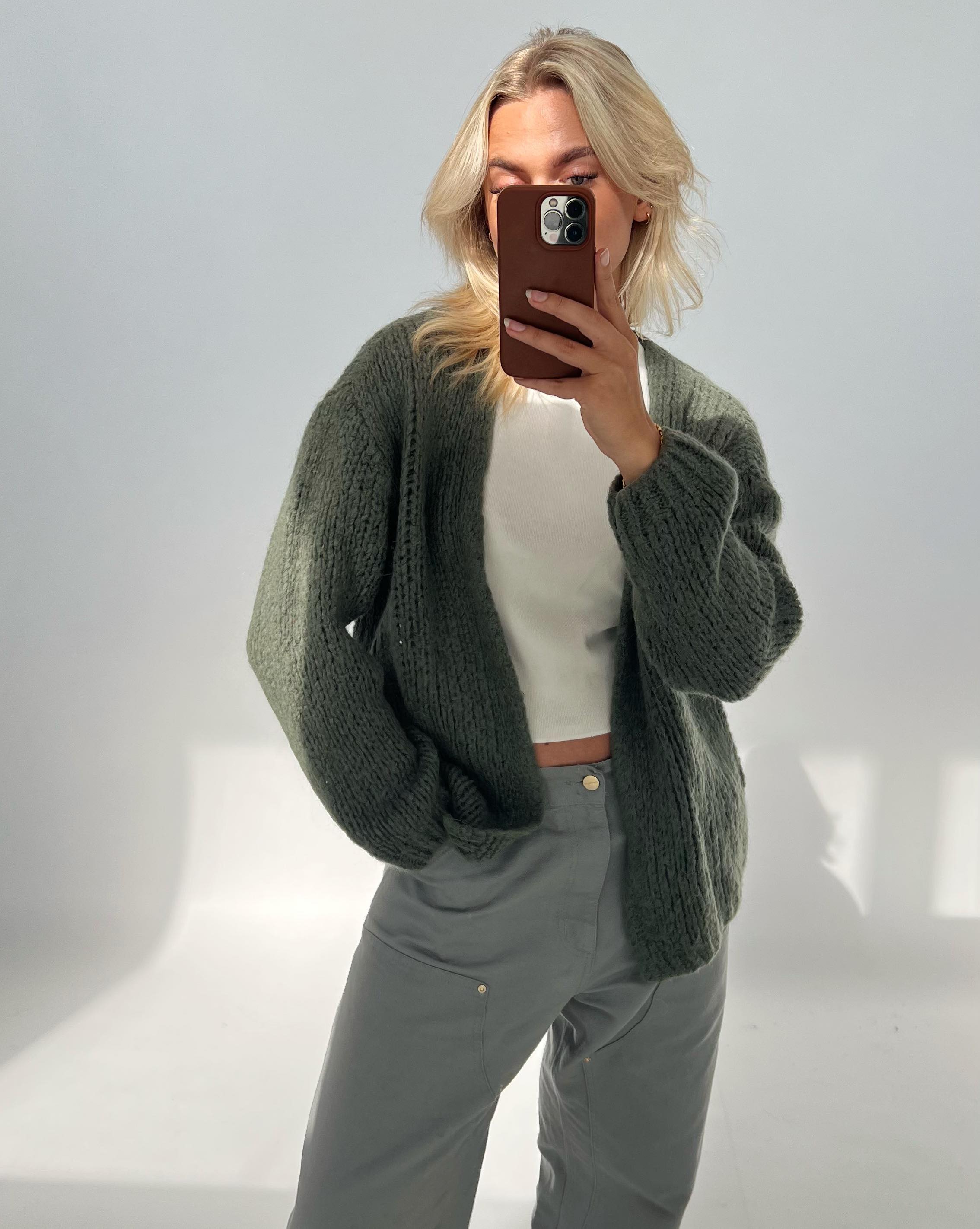 En person iført en grøn striktrøje og grå bukser tager en selfie med en telefon i et lyst rum med minimalistisk baggrund.