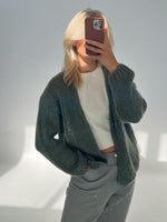 Person tager et billede med mobiltelefon, iført en striktrøje over en hvid T-shirt, stående i et rum med gråt sollys.