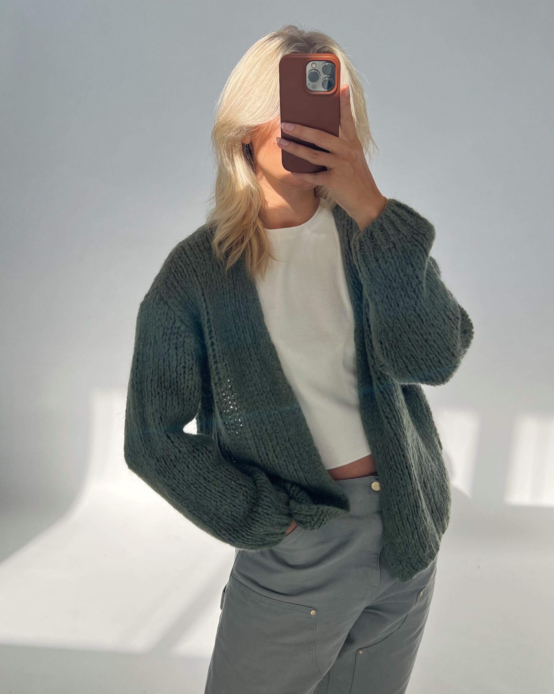 Person tager et billede med mobiltelefon, iført en striktrøje over en hvid T-shirt, stående i et rum med gråt sollys.