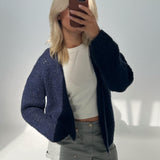 En person tager et spejlbillede og holder en mobiltelefon foran ansigtet. Personen har lyst hår, en mørk cardigan, en hvid trøje og grå bukser i et lyst rum.