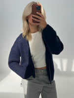 En person tager et spejlbillede og holder en mobiltelefon foran ansigtet. Personen har lyst hår, en mørk cardigan, en hvid trøje og grå bukser i et lyst rum.
