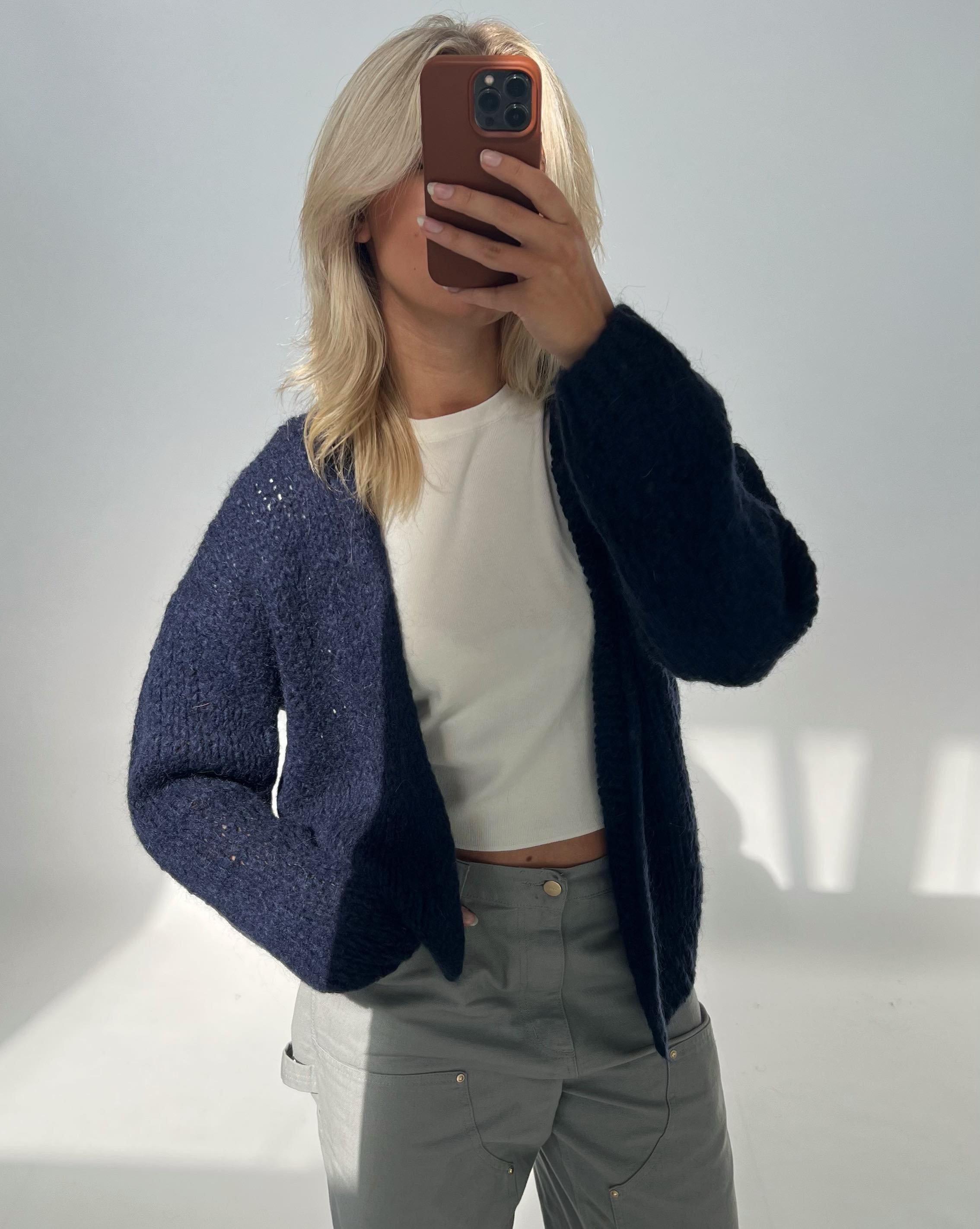 En person tager et spejlbillede og holder en mobiltelefon foran ansigtet. Personen har lyst hår, en mørk cardigan, en hvid trøje og grå bukser i et lyst rum.