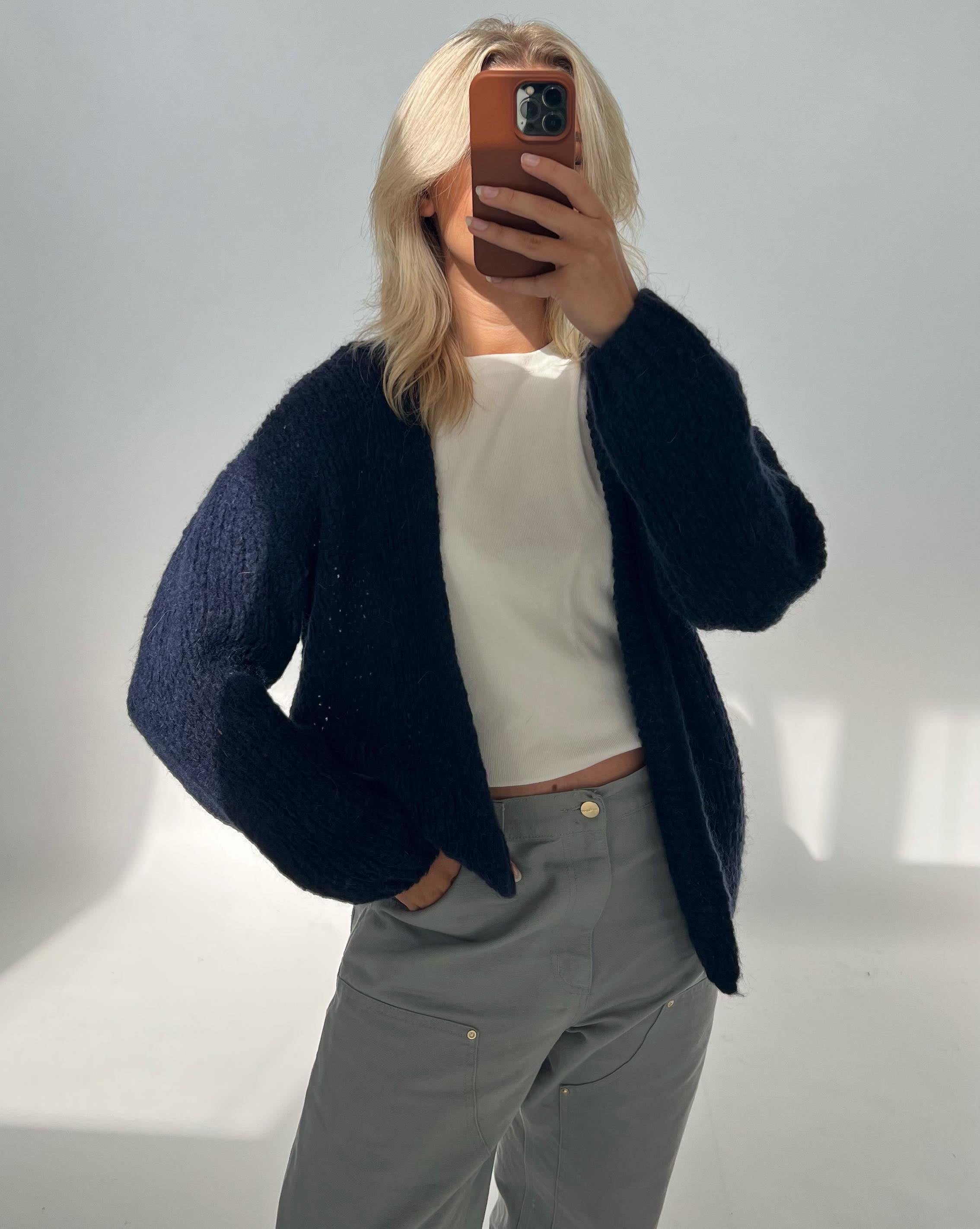 Person tager et spejlbillede med en smartphone, iført en mørk sweater og lyse bukser, i et minimalistisk, lyst rum uden tekst.