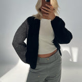 En person tager et spejlbillede iført en tyk sort sweater, hvid top og grå bukser, i et oplyst indendørs miljø. Personen skjuler ansigtet med en mobiltelefon.