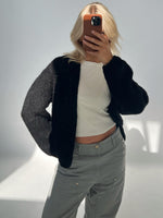 En person tager et spejlbillede iført en tyk sort sweater, hvid top og grå bukser, i et oplyst indendørs miljø. Personen skjuler ansigtet med en mobiltelefon.