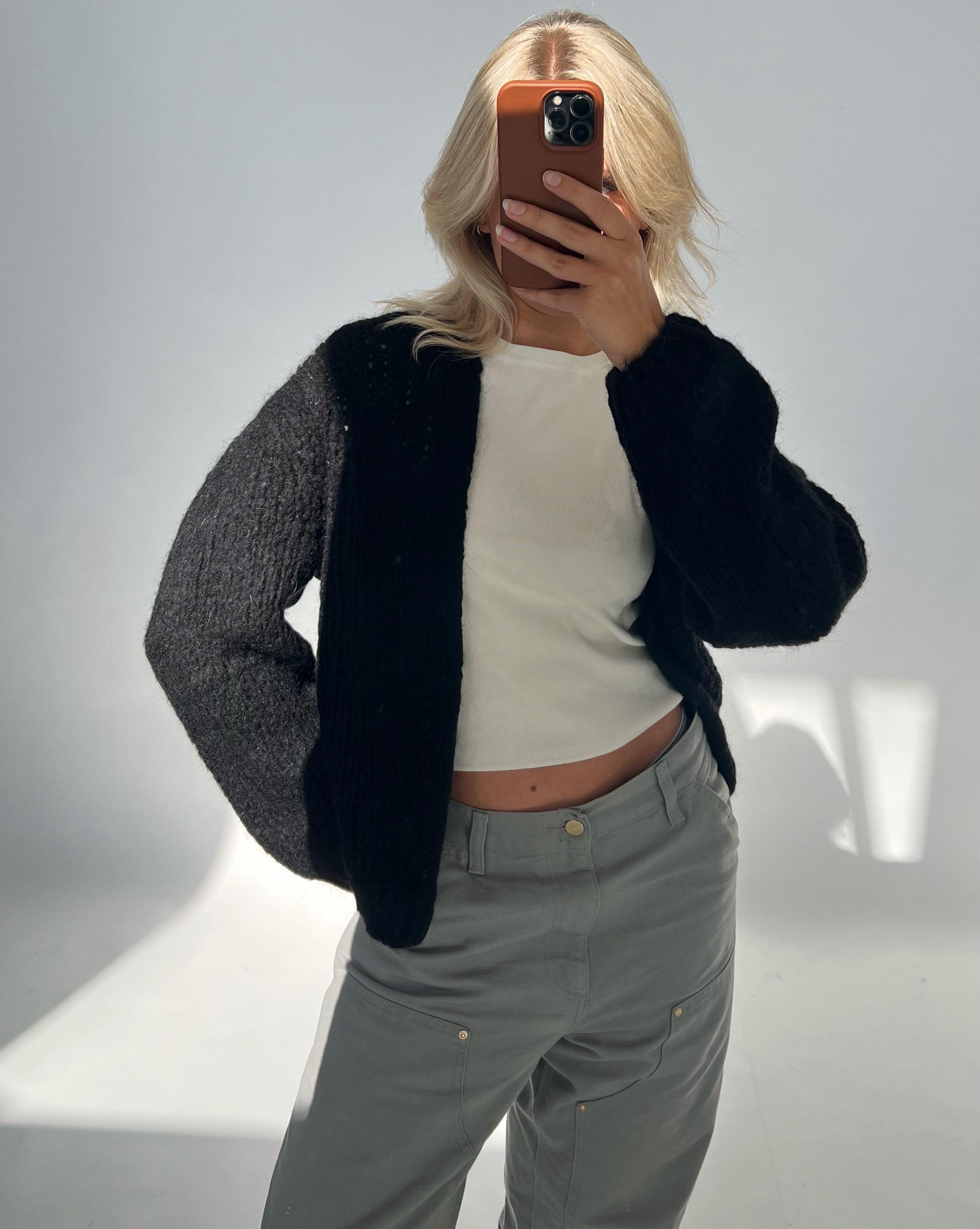 En person tager et spejlbillede iført en tyk sort sweater, hvid top og grå bukser, i et oplyst indendørs miljø. Personen skjuler ansigtet med en mobiltelefon.