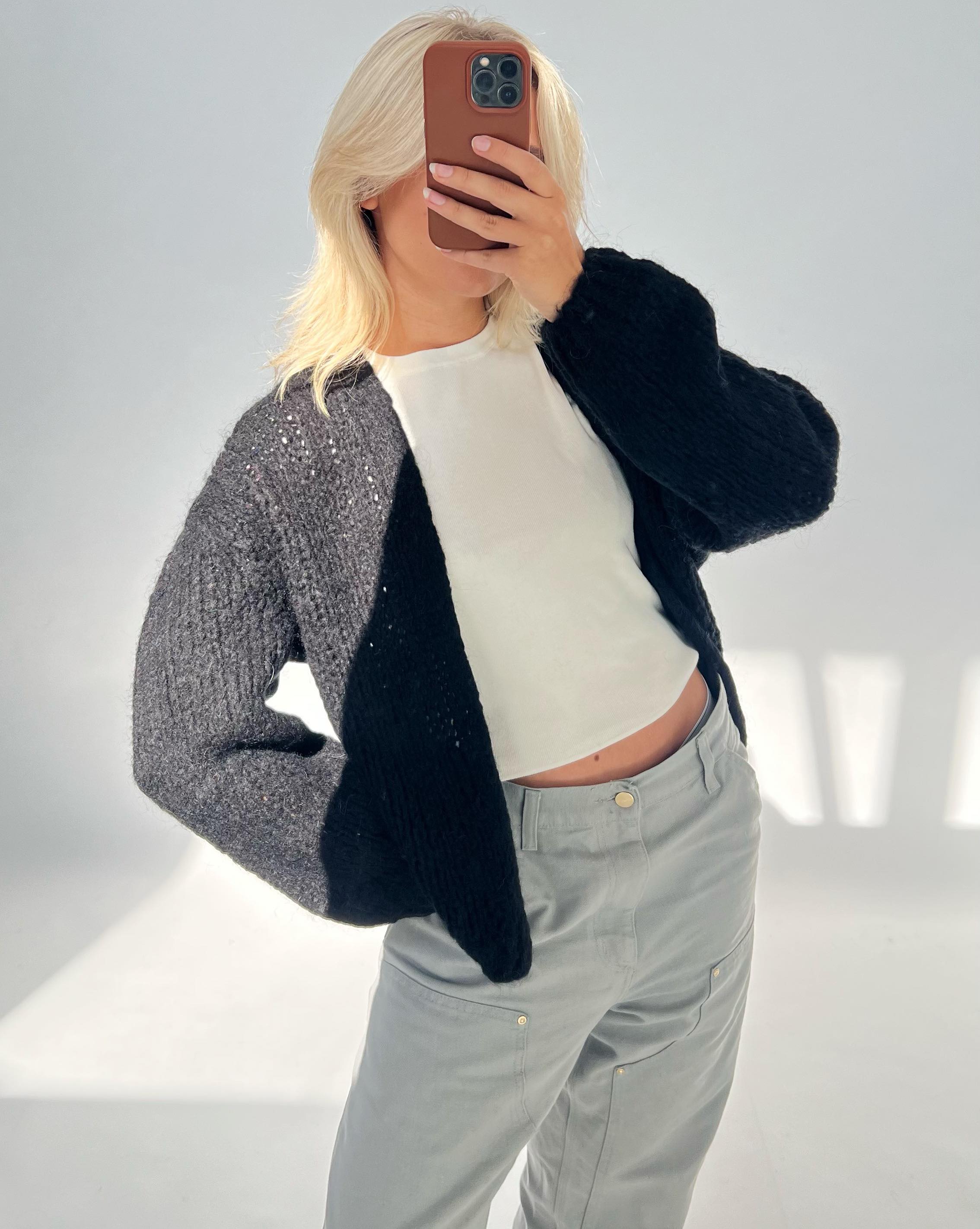 En person tager et spejlbillede med en mobiltelefon. De bærer en sort sweater og lysegrå bukser. Baggrunden er neutral og godt oplyst.