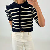 Kvinde iført sort-hvid-stribet cardigan med knapper, fotograferer sig selv med en smartphone. Hun har lyse jeans på og står i et neutralt, lyst rum.