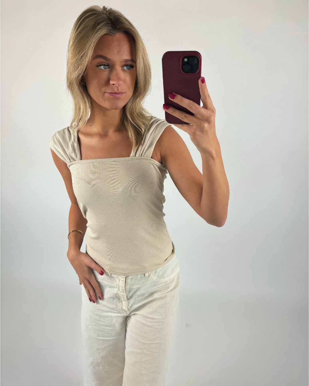En person tager en selfie med en smartphone. Personen bærer en beige top og hvide bukser. Baggrunden er en enkel, lys væg.