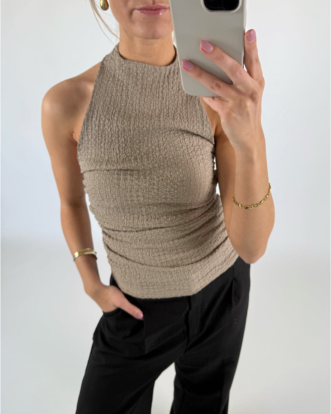 Kvinde tager et spejlbillede iført en beige halterneck-top og sorte bukser, med én hånd i lommen i et lyst rum. Hun bærer guldsmykker og holder en telefon.