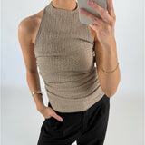 Kvinde tager et spejlbillede iført en beige halterneck-top og sorte bukser, med én hånd i lommen i et lyst rum. Hun bærer guldsmykker og holder en telefon.
