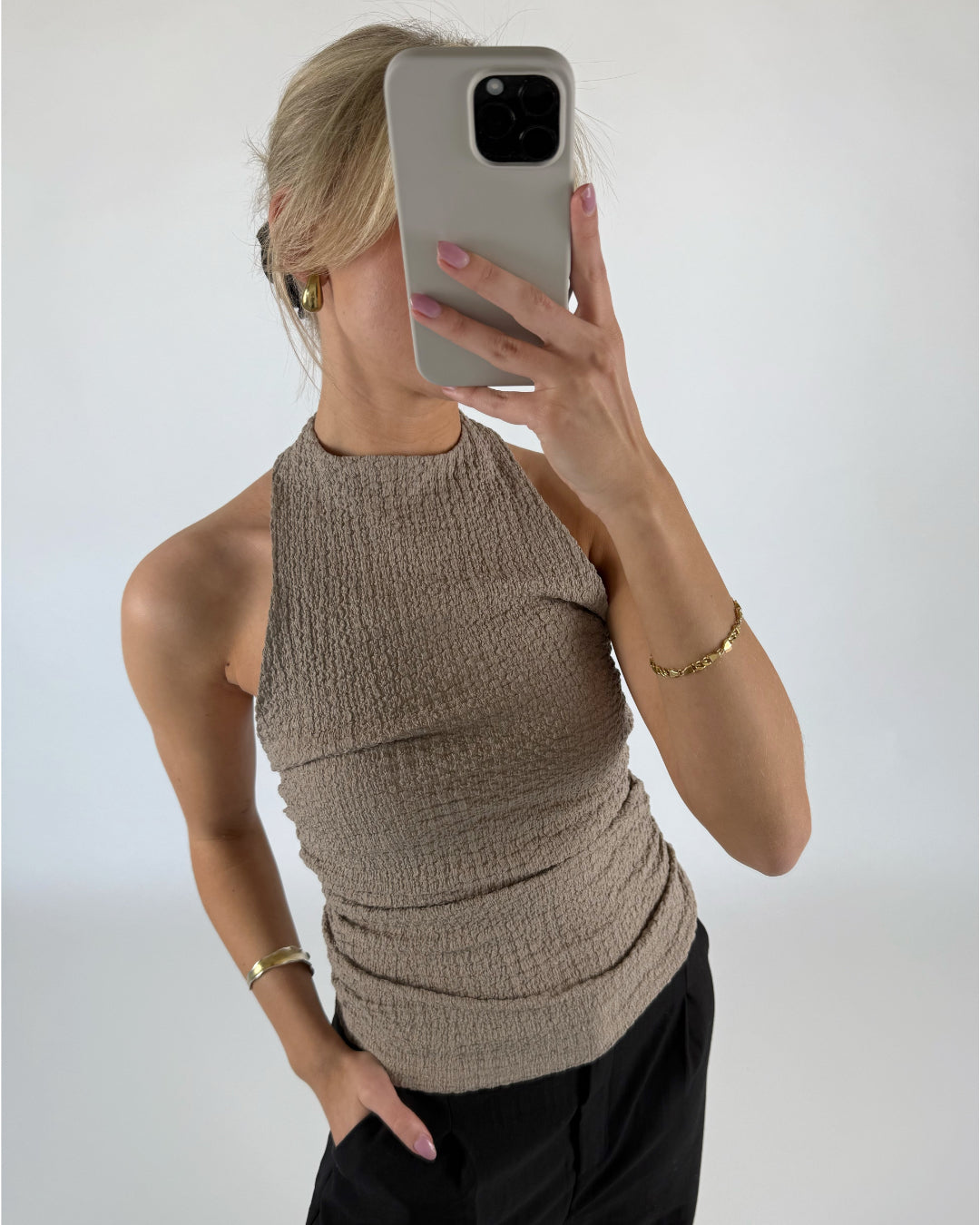 En person med beige top tager et spejlbillede med en mobiltelefon, iført guldøreringe og guldarmbånd. Personen står mod en lysegrå baggrund.