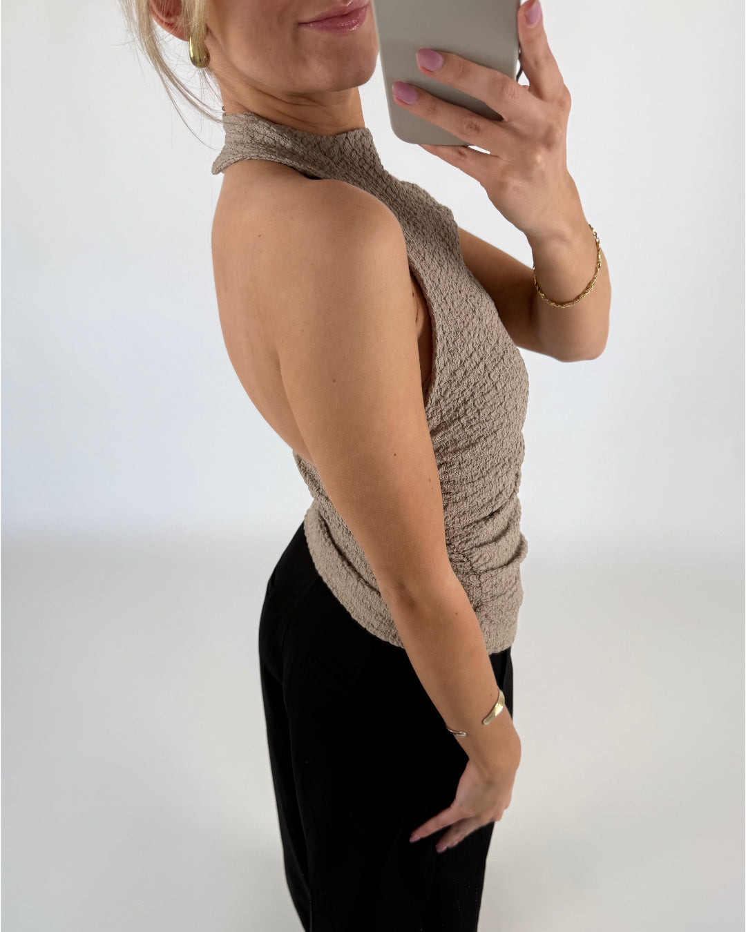 Kvinde iført beige halterneck-top, tager et spejlfoto med en smartphone. Hun har guldøreringe og armbånd, sort bukser, og befinder sig i et hvidt, minimalistisk rum.