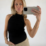 Kvinde tager en selfie med en smartphone, iført sort top og beige bukser. Hun poserer med et ansigtsudtryk og står i et lyst rum.