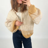 En person tager en selfie iført en strikket cardigan med beige og brune nuancer, stående i et neutralt rum. Ingen tekst til stede.