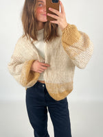 En person tager en selfie iført en strikket cardigan med beige og brune nuancer, stående i et neutralt rum. Ingen tekst til stede.