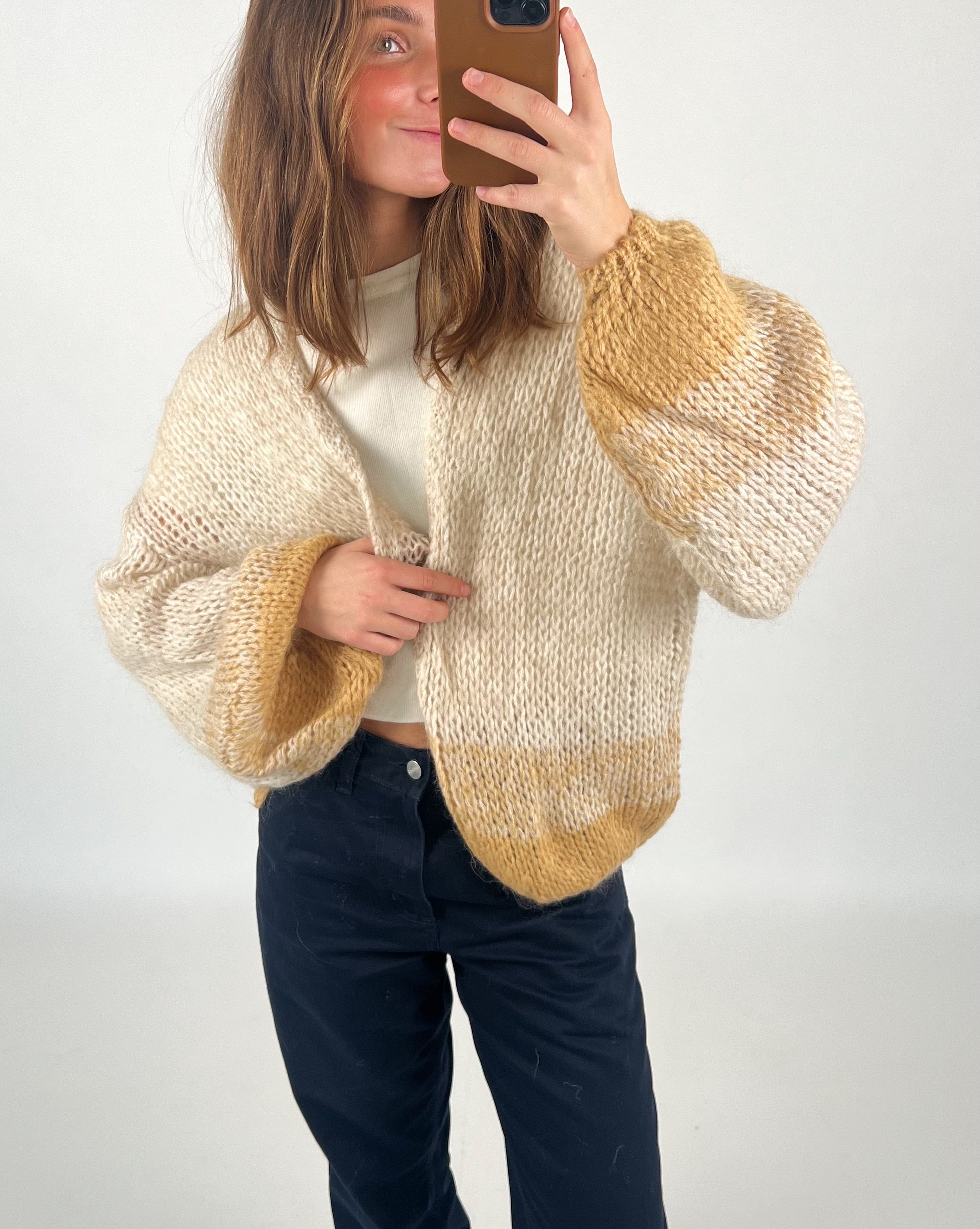 En person tager en selfie iført en strikket cardigan med beige og brune nuancer, stående i et neutralt rum. Ingen tekst til stede.