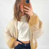 En person tager et spejlbillede iført en strikket cardigan med beige og guldfarvet mønster og en hvid t-shirt. Personen står i et enkelt, lyst rum.