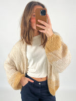 En person tager et spejlbillede iført en strikket cardigan med beige og guldfarvet mønster og en hvid t-shirt. Personen står i et enkelt, lyst rum.