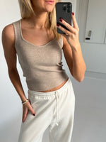 En person iført en beige tanktop tager en selfie med en smartphone i et lyst rum. Personen har hvide bukser og rød neglelak.
