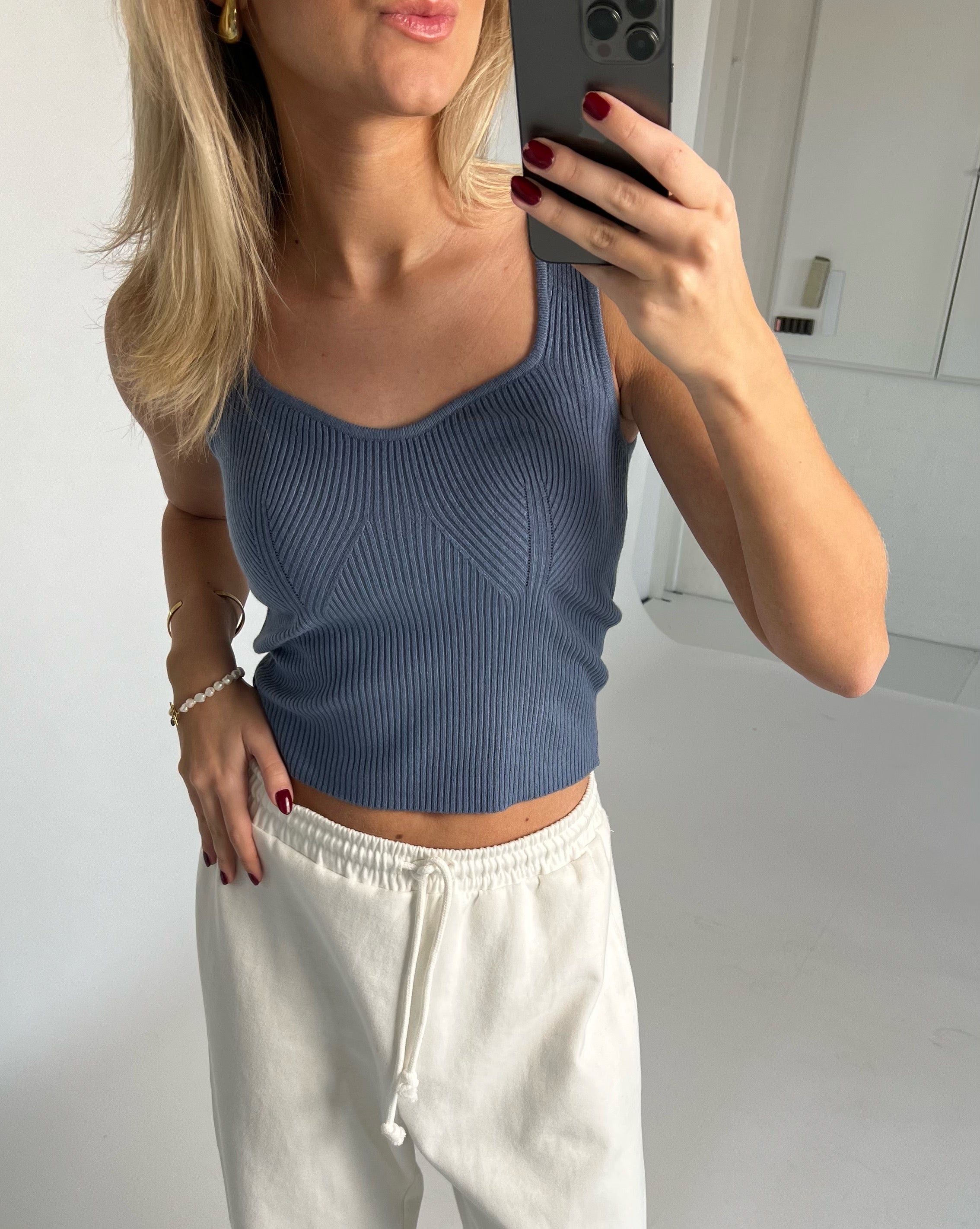 Kvinde tager selfie iført blå top og hvide bukser med snortræk i et lyst rum. Hendes hånd har røde negle og bærer smykker.