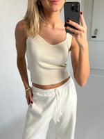 En person tager et spejlbillede iført en hvid top og joggebukser i et minimalistisk, lyst rum med en smartphone.