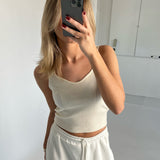 En person tager et spejlbillede med en telefon, iført en hvid tanktop og joggingbukser, i et minimalistisk rum med hvide vægge.