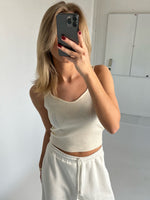 En person tager et spejlbillede med en telefon, iført en hvid tanktop og joggingbukser, i et minimalistisk rum med hvide vægge.