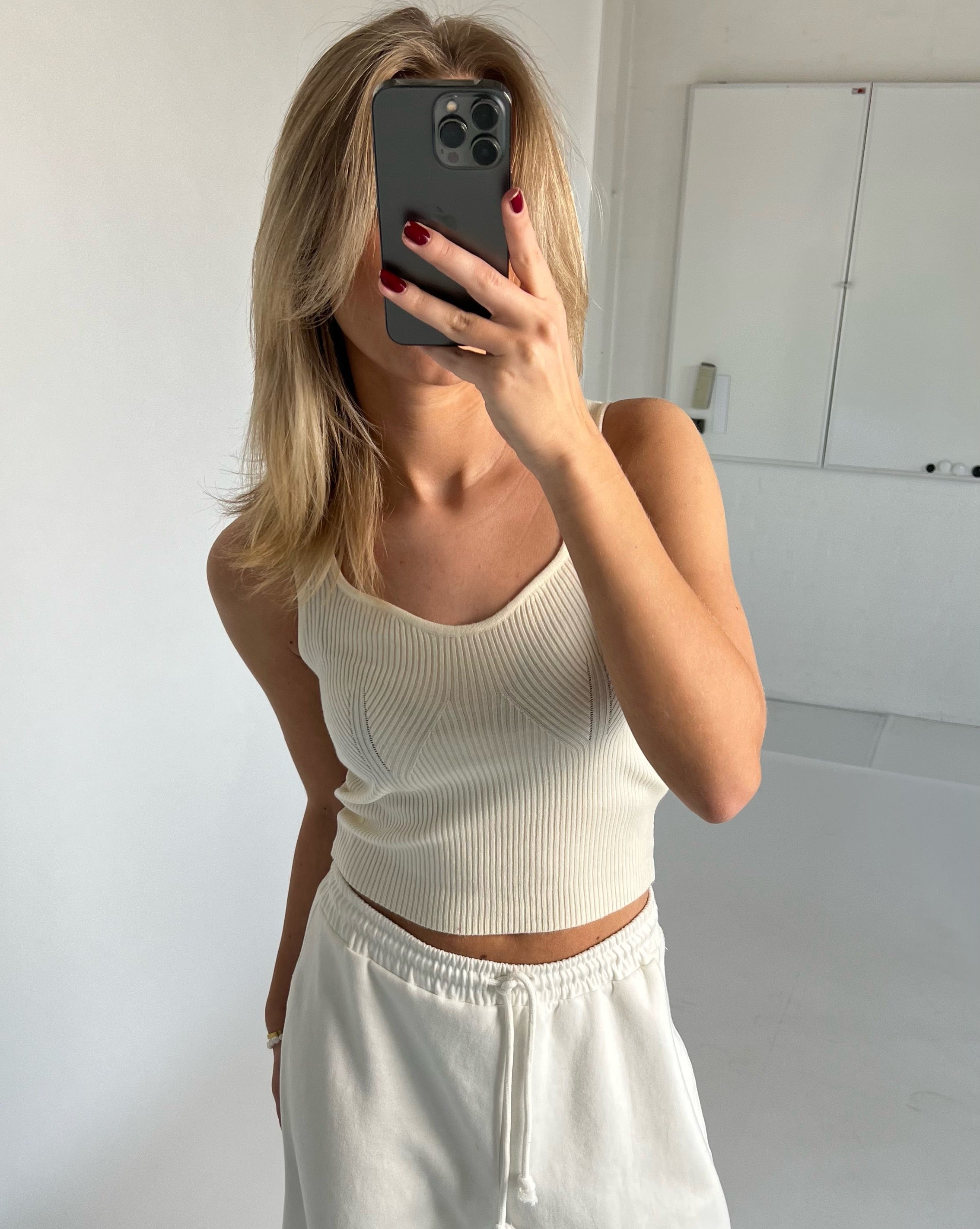 En person tager et spejlbillede med en telefon, iført en hvid tanktop og joggingbukser, i et minimalistisk rum med hvide vægge.